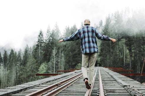Junger Mann balanciert auf Bahngleisen über eine Brücke im nebligen Wald - CAVF76966