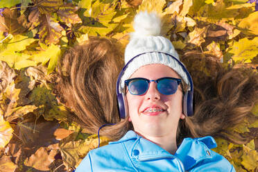 Teenager-Mädchen mit Sonnenbrille und Mütze liegt auf dem von gelben Blättern bedeckten Boden und genießt es, über Kopfhörer Musik zu hören. Das brünette Mädchen ist glücklich und lächelt. Horizonta - CAVF76981