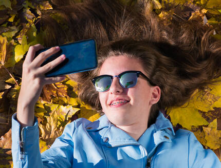 Teenager-Mädchen mit Sonnenbrille, das auf dem Boden liegt und von gelben Blättern bedeckt wird, während es mit seinem Handy ein Foto von sich macht. Das Mädchen lächelt in die Kamera. Horizontalaufnahme - CAVF76984