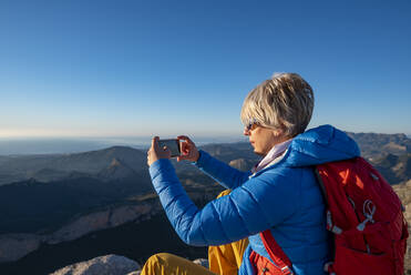 Junge Frau steht auf einem Felsen auf dem Gipfel eines Berges mit einem Mobiltelefon. - CAVF77028