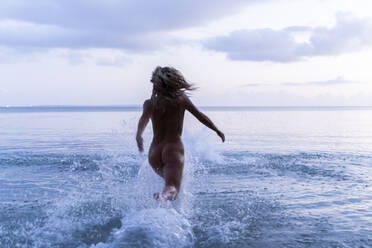 Back view of young woman running into the sea at sunset - AFVF05598