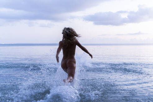 Rückenansicht einer jungen Frau, die bei Sonnenuntergang ins Meer läuft - AFVF05598