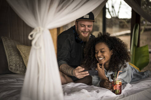 Glückliches Paar auf dem Bett in einer Lodge mit Smartphone und frischem Eistee - MJRF00263