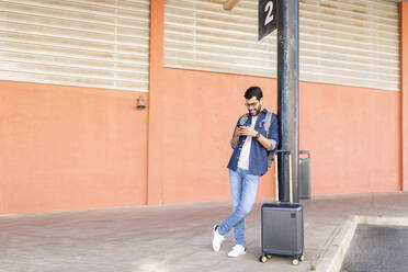 Mann mit Rucksack und Trolley-Tasche wartet an der Bushaltestelle und benutzt ein Mobiltelefon - JPTF00464