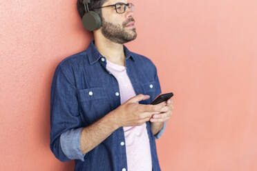 Man with smartphone leaning against pink wall listening music with wireless headphones - JPTF00469