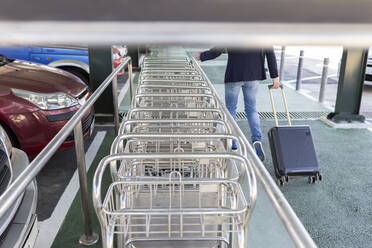 Crop view of man pulling trolley bag walking along row of baggage carts - JPTF00477