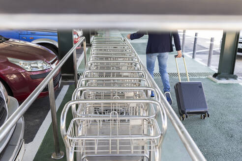 Crop-Ansicht von Mann zieht Trolley Tasche zu Fuß entlang Reihe von Gepäckwagen - JPTF00477