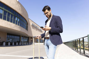 Porträt eines Geschäftsmannes mit Trolley-Tasche, der vor einem Flughafen steht und auf sein Handy schaut - JPTF00488