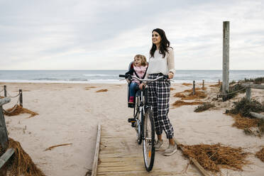 Woman with bicycle on a boardwalk in the dunes with daughter in child's seat - JRFF04136