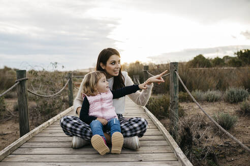Happy mother and daughter sitting on a boardwalk in the countryside - JRFF04162