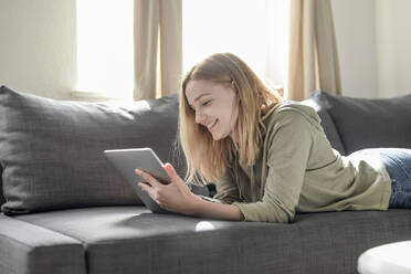 Porträt einer lächelnden jungen Frau, die zu Hause auf der Couch liegt und ein digitales Tablet benutzt - BFRF02197