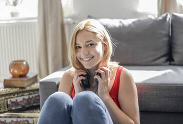 Portrait of happy young woman sitting on the floor at home listening music with headphones - BFRF02201