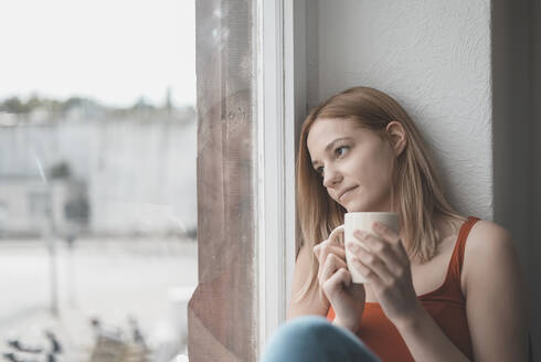 Porträt einer nachdenklichen jungen Frau mit einer Tasse Kaffee, die aus dem Fenster schaut - BFRF02203