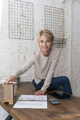 Portrait of smiling mature woman sitting on desk in architectural office - AFVF05639