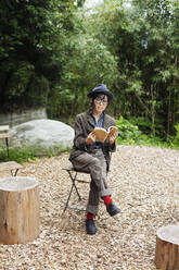 Japanerin mit Brille und Hut sitzt auf einem Stuhl vor einem Öko-Café und liest ein Buch. - MINF14069