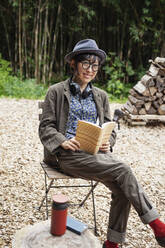 Japanese woman wearing glasses and hat sitting on chair outside Eco Cafe, reading book. - MINF14072