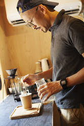 Japanischer Mann mit Baseballkappe und Brille in einem Öko-Café, der eine Tasse Kaffee zubereitet. - MINF14076