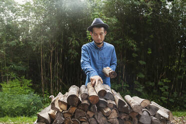 Japanese man wearing hat standing outdoors, stacking logs of firewood. - MINF14095