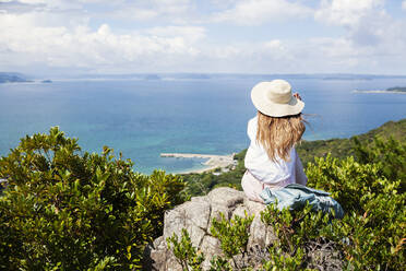Japanische Frau mit Hut, die auf einem Felsen auf einer Klippe sitzt, das Meer im Hintergrund. - MINF14099