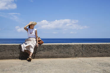 Japanische Frau mit Hut, die auf einer Mauer sitzt, im Hintergrund das Meer. - MINF14109