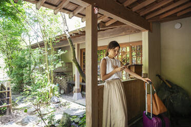 Japanische Frau steht auf einer Veranda, benutzt ein Mobiltelefon, hält einen Koffer und eine Handtasche. - MINF14111