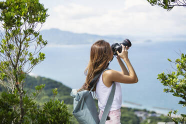 Japanische Frau mit Rucksack fotografiert auf einer Klippe, das Meer im Hintergrund. - MINF14127