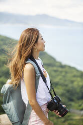 Japanische Frau mit Rucksack und Kamera in der Hand steht auf einer Klippe, im Hintergrund das Meer. - MINF14128