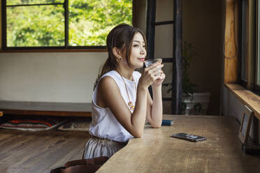 Smiling Japanese woman sitting at a table in a Japanese restaurant, drinking. - MINF14132