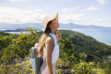 Japanische Frau mit Hut und Rucksack auf einer Klippe stehend, das Meer im Hintergrund. - MINF14171