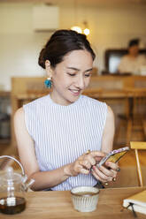 Japanische Frau sitzt an einem Tisch in einem vegetarischen Café und benutzt ein Mobiltelefon. - MINF14206