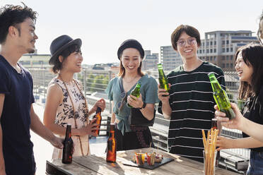Eine Gruppe junger japanischer Männer und Frauen steht auf einem Dach in einer städtischen Umgebung und trinkt Bier. - MINF14234
