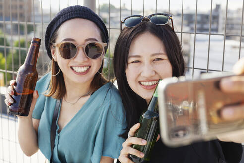 Zwei junge Japanerinnen sitzen auf einem Dach in einer städtischen Umgebung und machen ein Selfie mit ihrem Mobiltelefon. - MINF14239