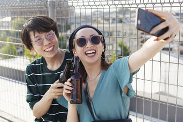 Young Japanese man and woman sitting on a rooftop in an urban setting, taking selfie with mobile phone. - MINF14240