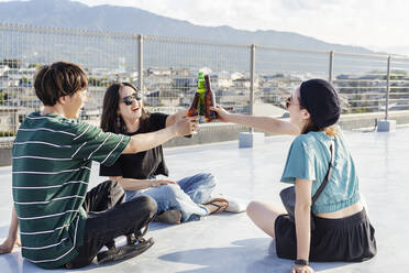 Ein junger japanischer Mann und zwei Frauen sitzen auf einem Dach in einer städtischen Umgebung und trinken Bier. - MINF14243