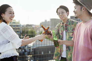 Junge japanische Männer und Frauen stehen auf einem Hausdach in einer städtischen Umgebung und trinken Bier. - MINF14248