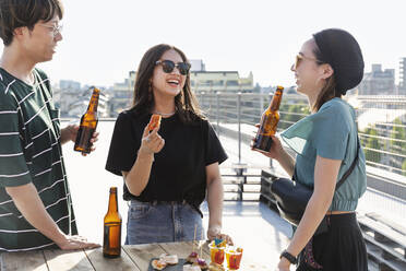 Ein junger japanischer Mann und zwei Frauen stehen auf einem Dach in einer städtischen Umgebung und trinken Bier. - MINF14260