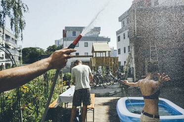 Cropped Hand hält Rohr mit Wasser spritzt auf Frau von Gebäuden gegen den Himmel - EYF00142