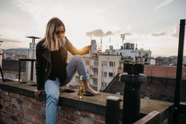 Frau mit Sonnenbrille und Getränk auf einer Stützmauer in der Stadt sitzend - EYF00344