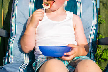 Mittelteil eines Jungen, der einen Lutscher isst, während er auf einem Stuhl im Hof sitzt - EYF00412