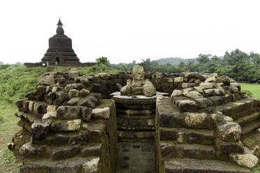 Eine zerstörte Steinstupa mit beschädigter Buddha-Statue in der Mitte, mit der West Myatazaung Pagode im Hintergrund, Mrauk U, Rakhine, Myanmar (Burma), Asien - RHPLF14058