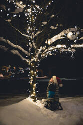 Junge mit Weihnachtsmannmütze sitzt im Schnee und betrachtet die Weihnachtsbeleuchtung am Baum. - CAVF77088