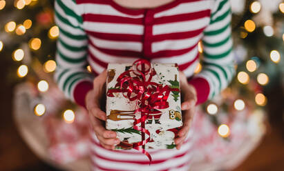 Kind im Schlafanzug mit Weihnachtsgeschenk vor Lichterketten am Baum. - CAVF77115