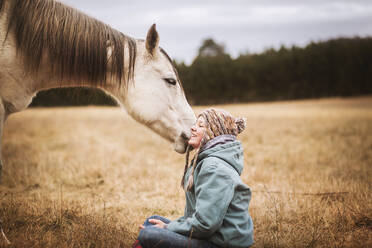 Weißes Pferd zerrt an Mädchen Hut im Feld im Herbst, während Mädchen sitzt - CAVF77129