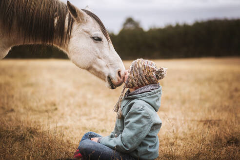 Junges Mädchen küsst weißes Pferd auf die Nase, während sie auf dem Boden sitzt - CAVF77130