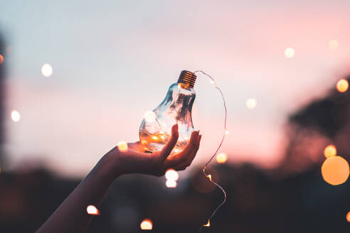 Close-Up Of Woman Holding Illuminated Light Bulb Against Sky At Night - EYF00690