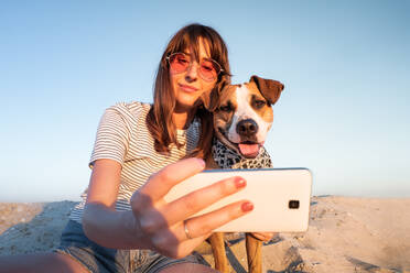 Low Angle View Of Young Woman Taking Selfie With Dog Through Smart Phone At Beach Against Clear Sky - EYF00805