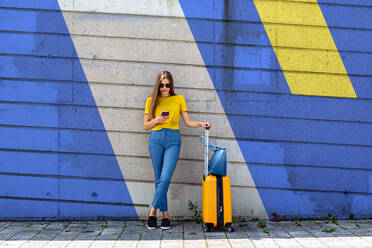 Junge Frau mit Gepäck, die ein Mobiltelefon benutzt, während sie an der Wand steht - EYF00972