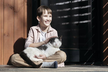 Smiling boy looking away while stroking cat relaxing on him at porch during sunny day, Tarusa, Russia - EYAF00951