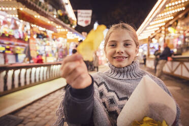 Portrait of smiling girl eating potato chip at carnival in city during night. Munich, Germany - DHEF00148