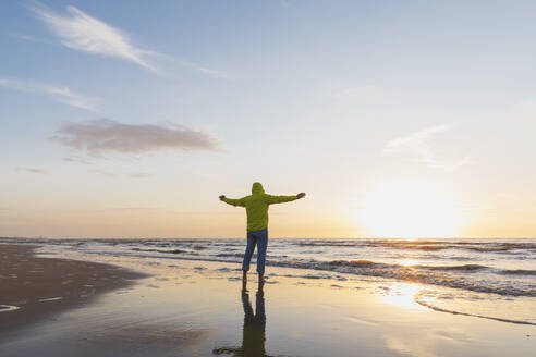 Älterer Mann mit ausgestreckten Armen, der bei Sonnenuntergang am Strand steht, Nordseeküste, Flandern, Belgien - GWF06543